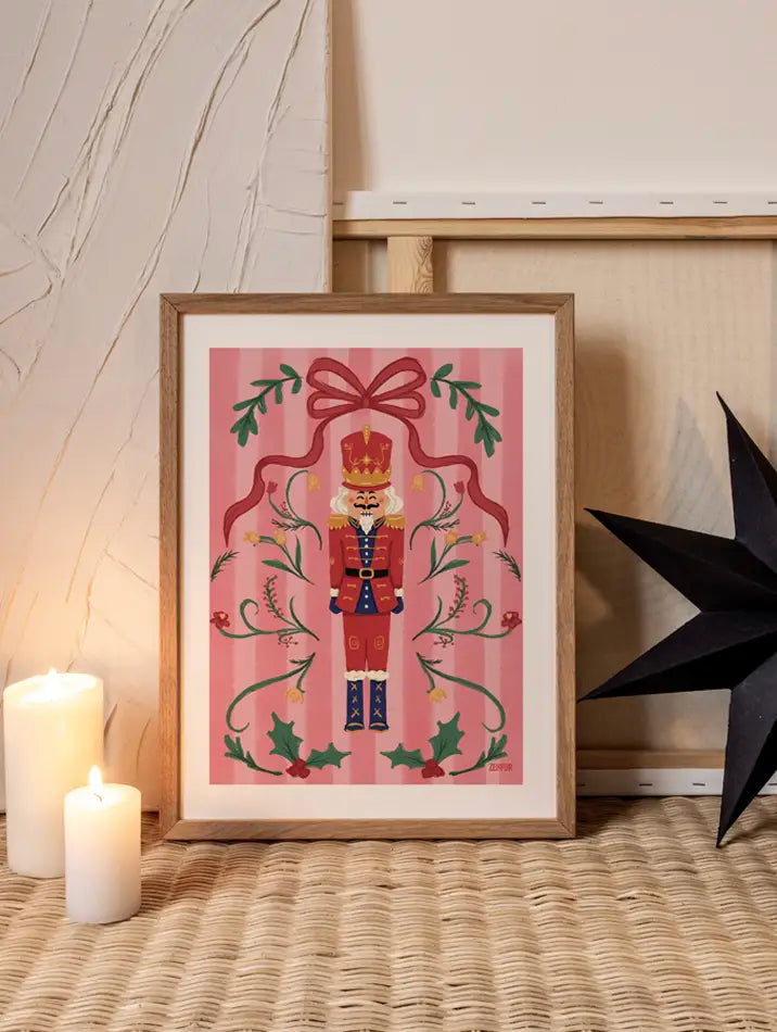 Poster artistique Casse-Noisette illustré par Blanca Flor, représentant un soldat de Noël entouré de houx, fleurs et un grand nœud rouge, parfait comme tableau pour une ambiance festive élégante sur un fond rose pastel.